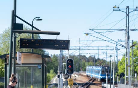Perrongmiljö vid Roslagsbanan med tåg, skyltning och resenärer.